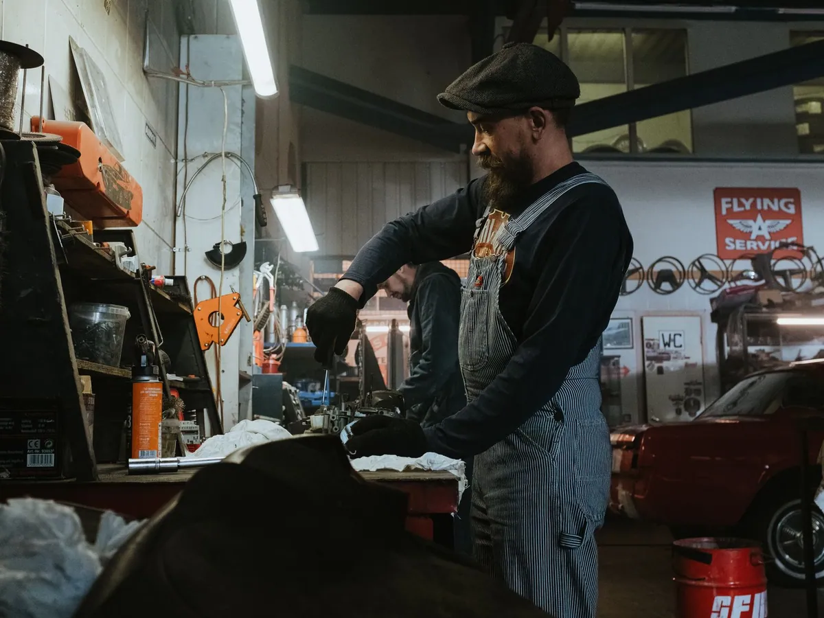 Mechanic performing repairs in a vintage garage setting, using tools and machinery.